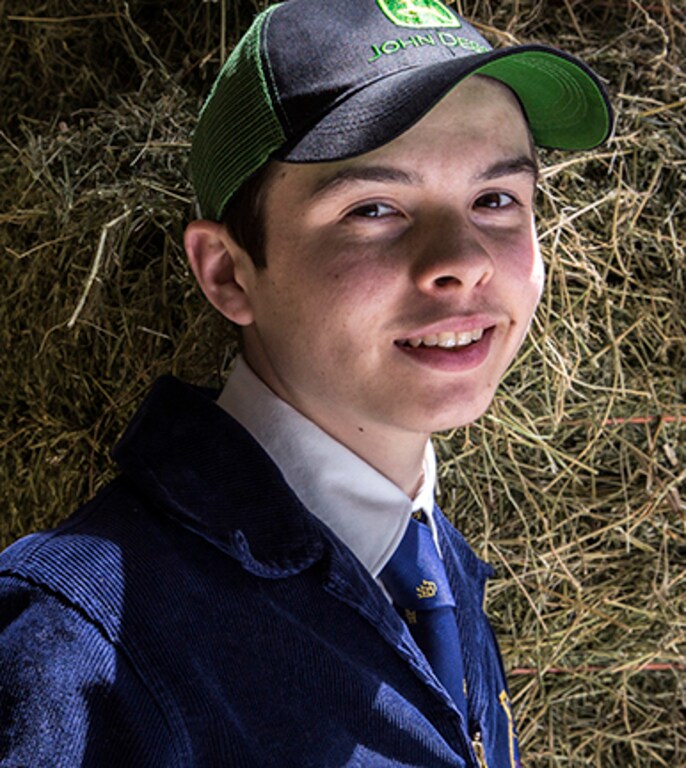 An FFA student wearing a John Deere hat stands in front of a hay bale