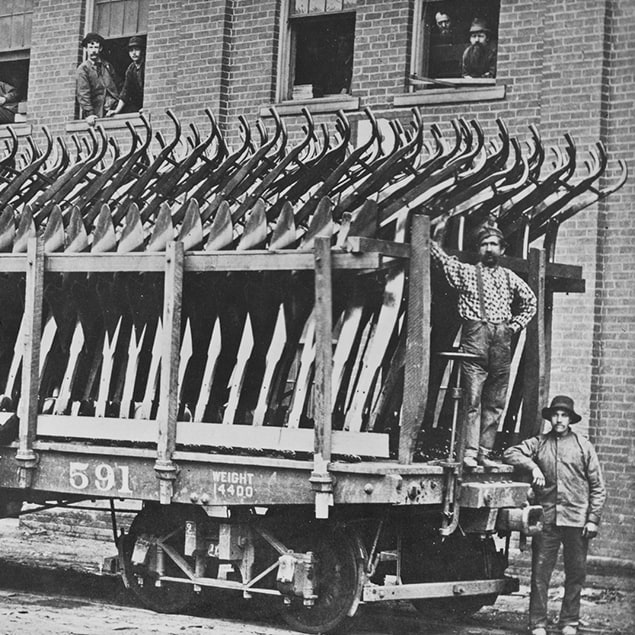 Photographie de 1882 illustrant trois hommes se tenant à côté du wagon ferroviaire Deere & Co chargé de charrues en acier prêtes à être expédiées, ainsi que les employés de l’usine regardant par la fenêtre du bâtiment situé derrière
