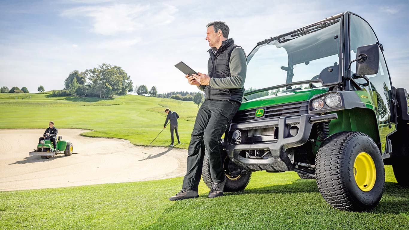 Ein Head Greenkeeper prüft die Daten auf seinem Tablet, während zwei Greenkeeper im Hintergrund auf einem Golfplatz arbeiten.
