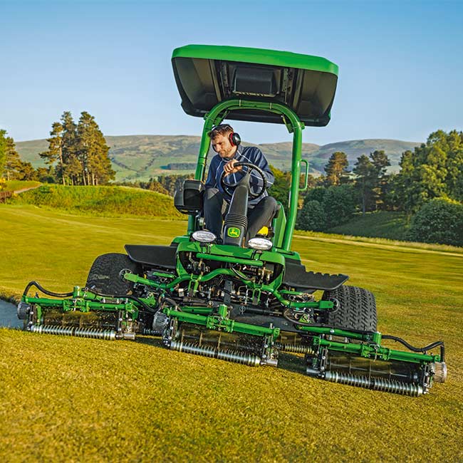 Ein Greenkeeper arbeitet auf einer Rasenfläche mit einem John Deere Greens-Mäher in Frontansicht.