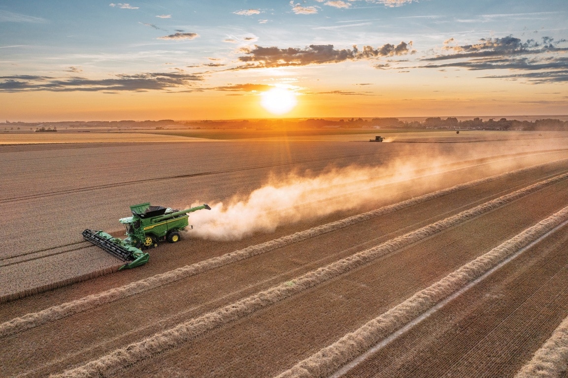 Une moissonneuse-batteuse en train de récolter du blé au coucher du soleil, laissant un long sillage de poussière derrière elle, à travers un vaste paysage agricole