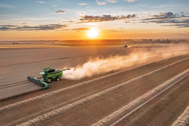 Une moissonneuse-batteuse en train de récolter du blé au coucher du soleil, laissant un long sillage de poussière derrière elle, à travers un vaste paysage agricole