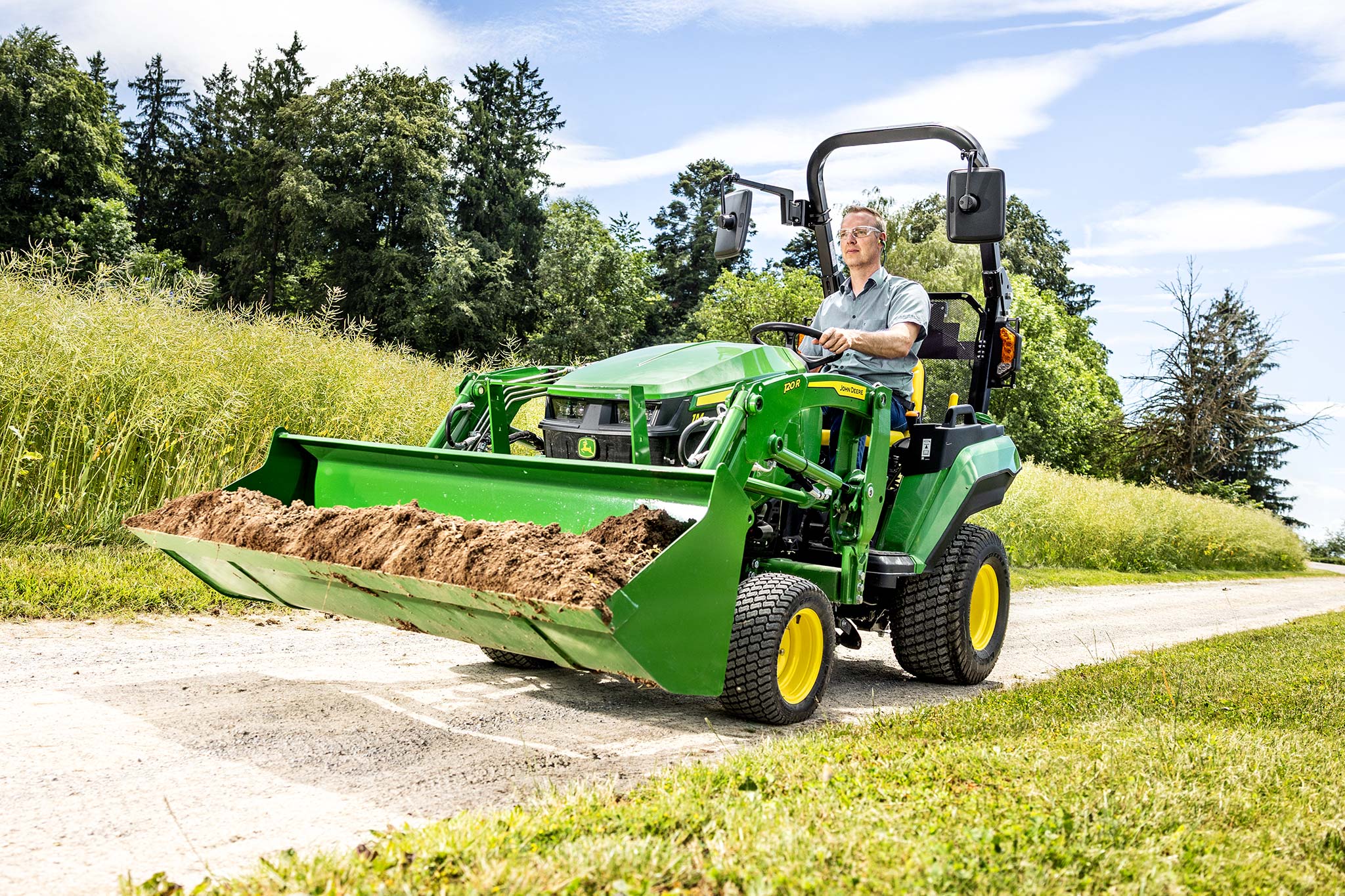 Fahrer, der einen John Deere Kompakttraktor fährt, der Erde in einer Frontladerschaufel auf einer Landstraße transportiert