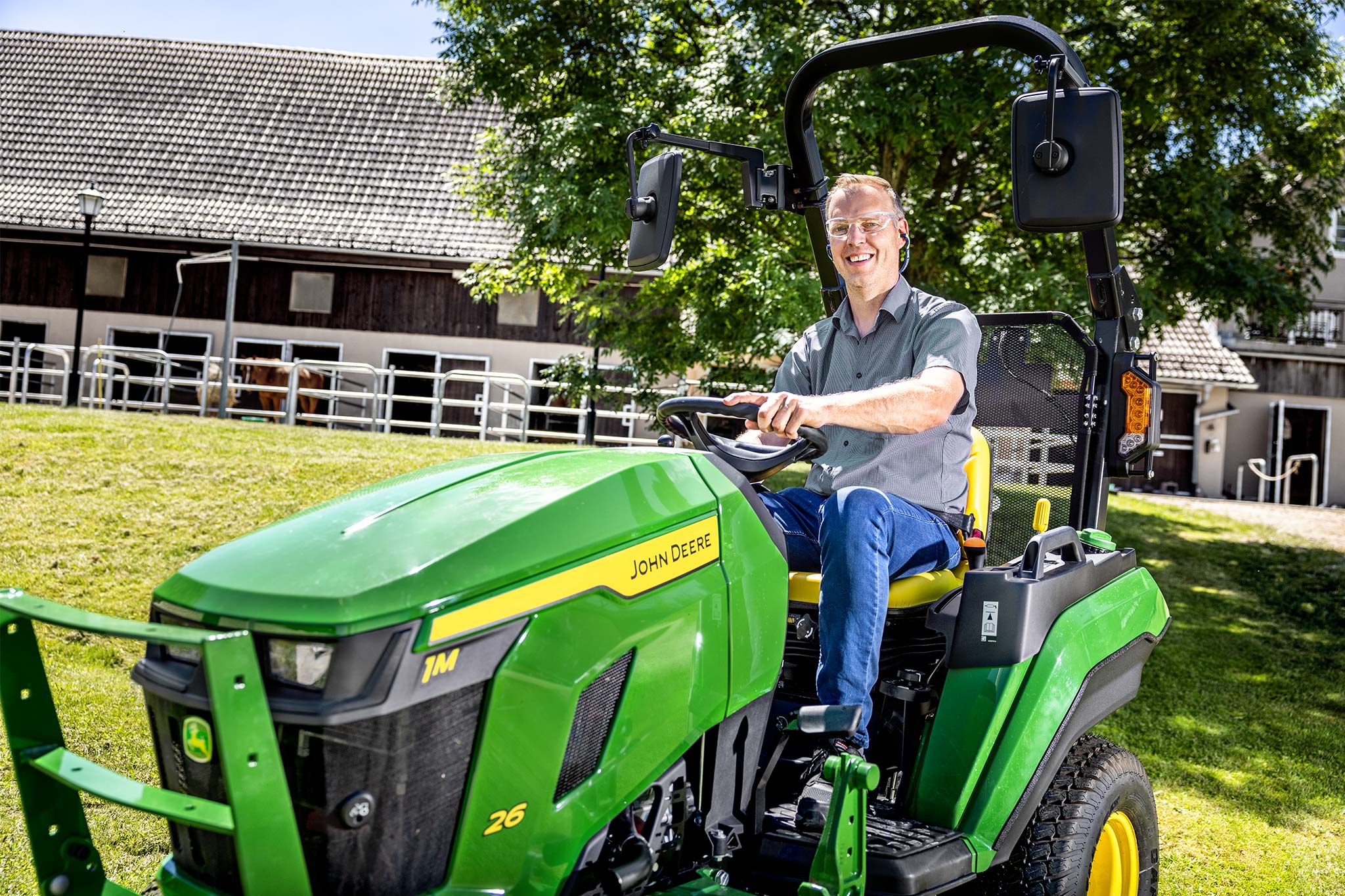 Fahrer, der einen John Deere Kompakttraktor im Freien in der Nähe von Wirtschaftsgebäuden und eingezäunten Weideflächen fährt
