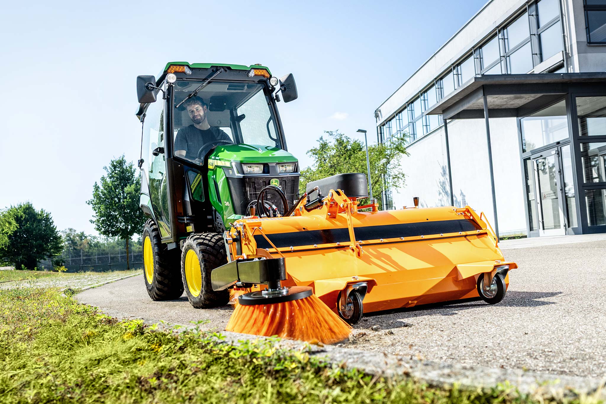 John Deere Kompakttraktor mit geschlossener Kabine und frontseitig montierter Kehrmaschine zur Reinigung eines gepflasterten Bereichs