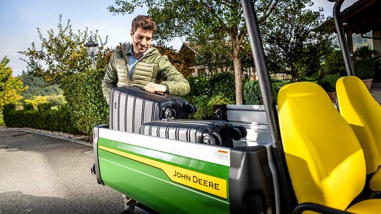 Une personne charge une grande valise noire dans le coffre d’un Gator&nbsp;GS John&nbsp;Deere équipé de sièges jaune vif, entouré de verdure et d’arbres.