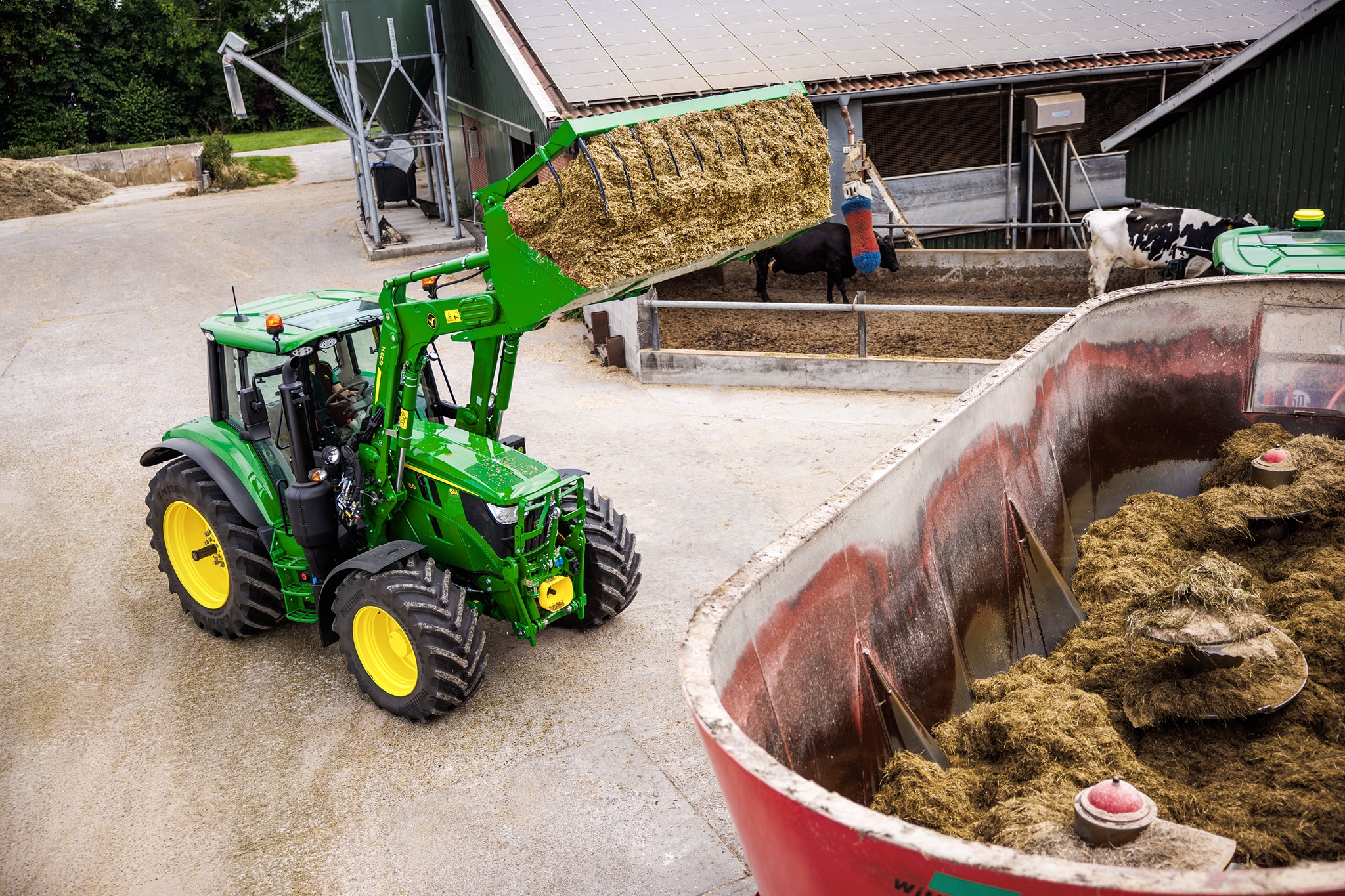 John Deere Traktoren der Serie 6M mit einem Frontlader zur Zuführung von Silage in einen Mischwagen auf einem Bauernhof.