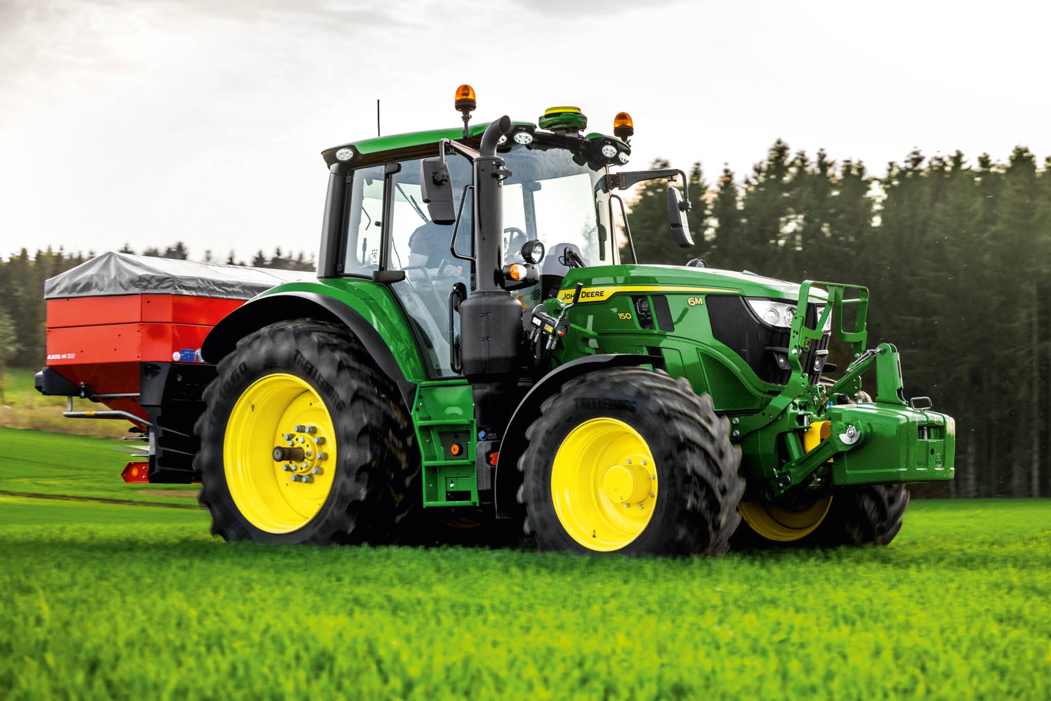 John Deere Traktor der Serie 6M mit heckmontiertem Streuer, der auf Gras in der Nähe eines Waldrands betrieben wird.