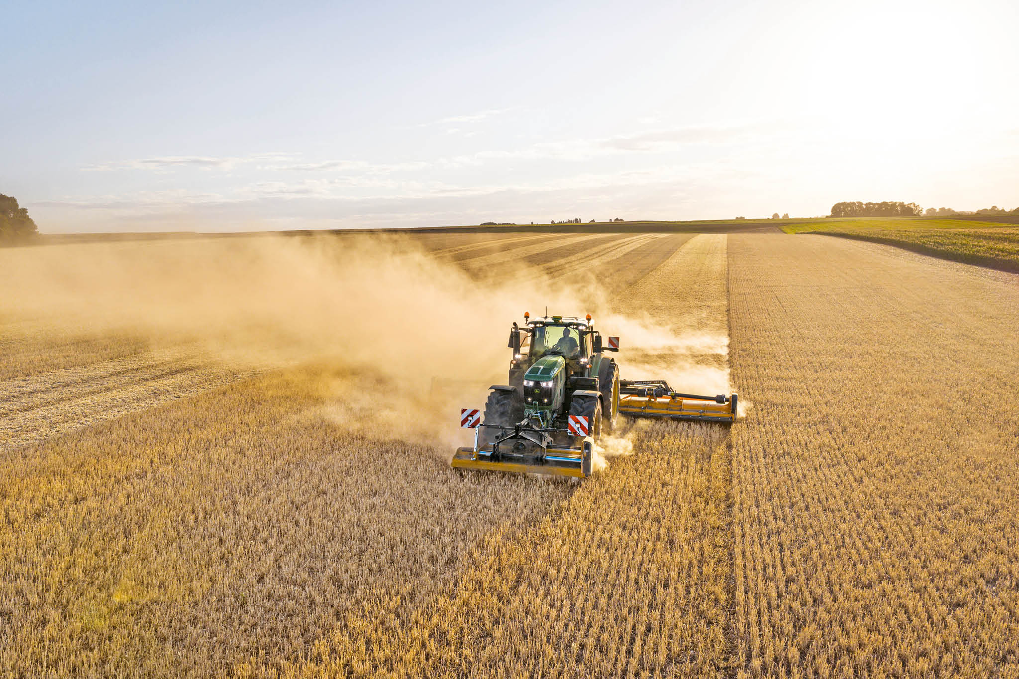 Un tracteur John&nbsp;Deere de la série&nbsp;6M tractant un épandeur à lisier sur une route de campagne bordée d’herbe et de champs.