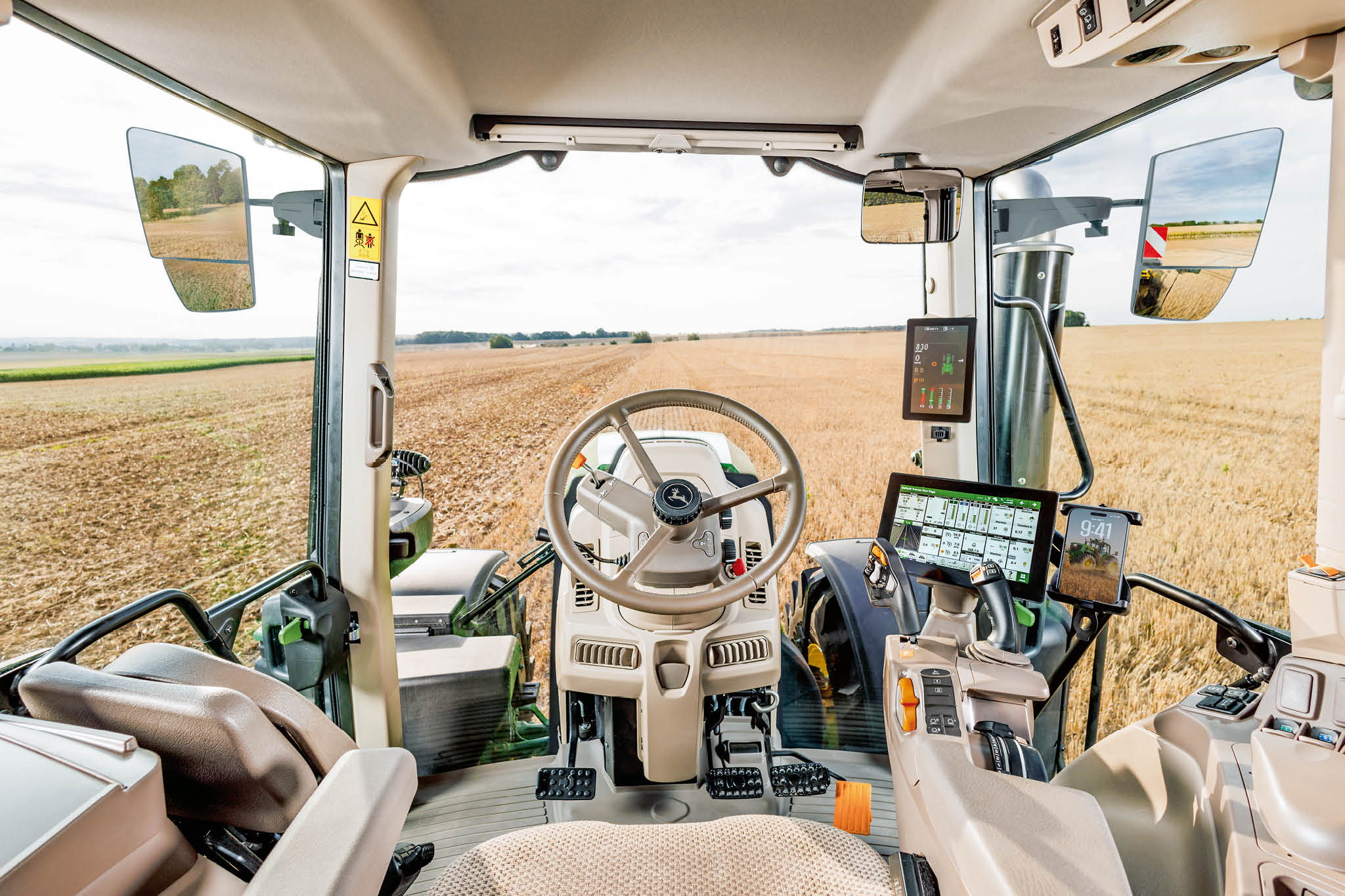 Vue intérieure de la cabine d’un tracteur John&nbsp;Deere de la série 6M, montrant le volant, les consoles de commande et un champ de chaume devant.