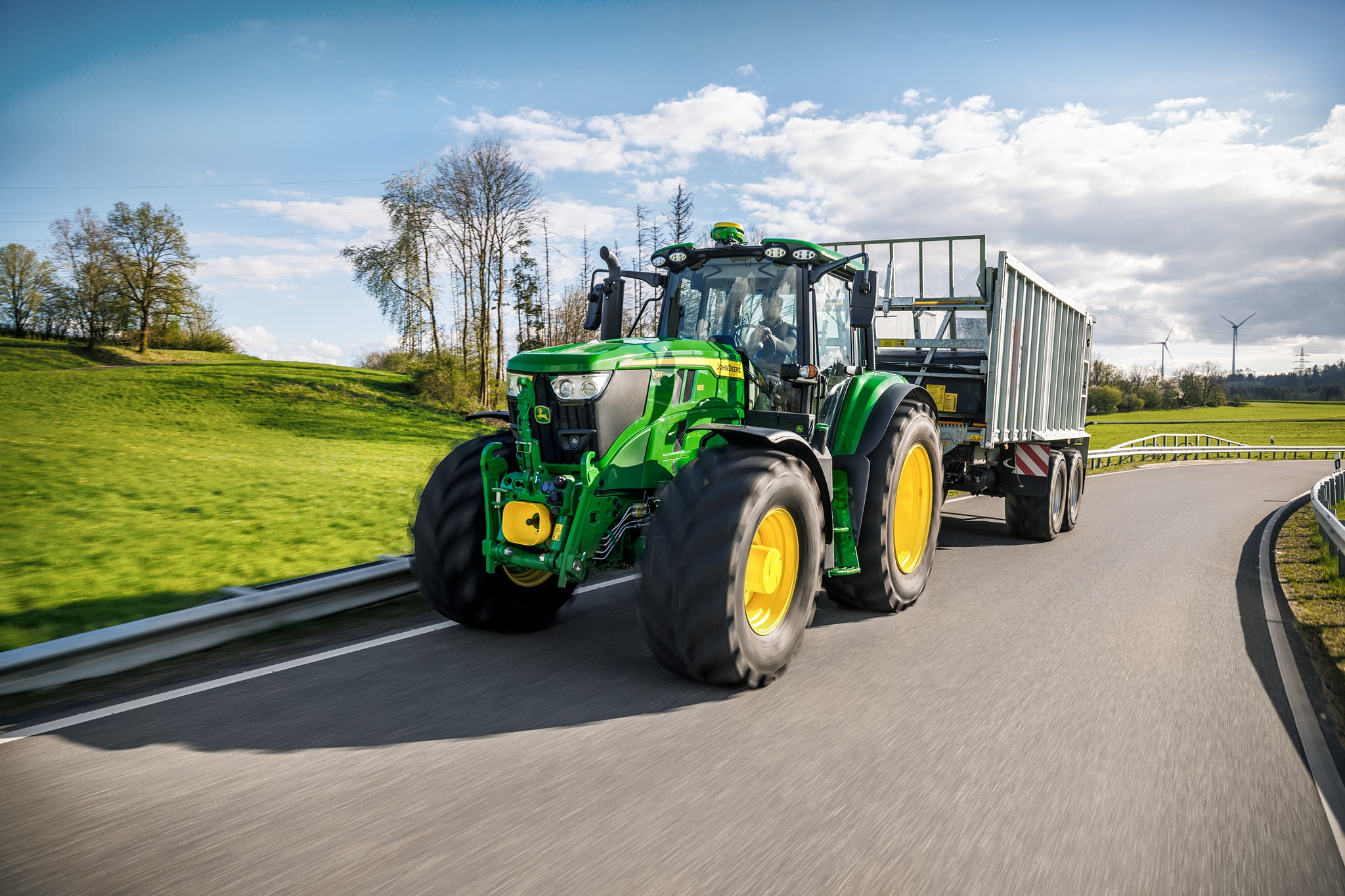 John Deere Traktoren der Serie 6M transportieren einen hochseitigen Anhänger auf einer asphaltierten Landstraße.