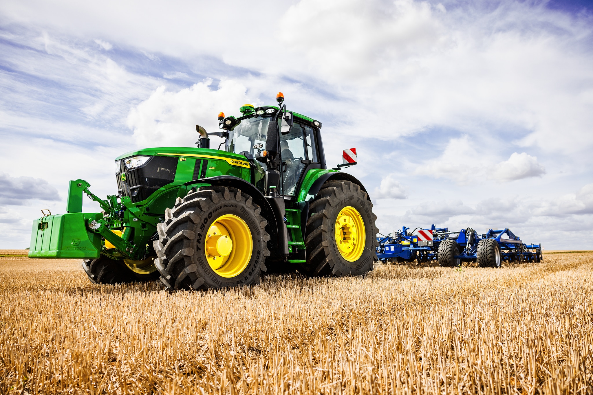 John Deere Traktoren der Serie 6M ziehen ein schweres Bodenbearbeitungsgerät unter bewölktem Himmel über ein Stoppelfeld.