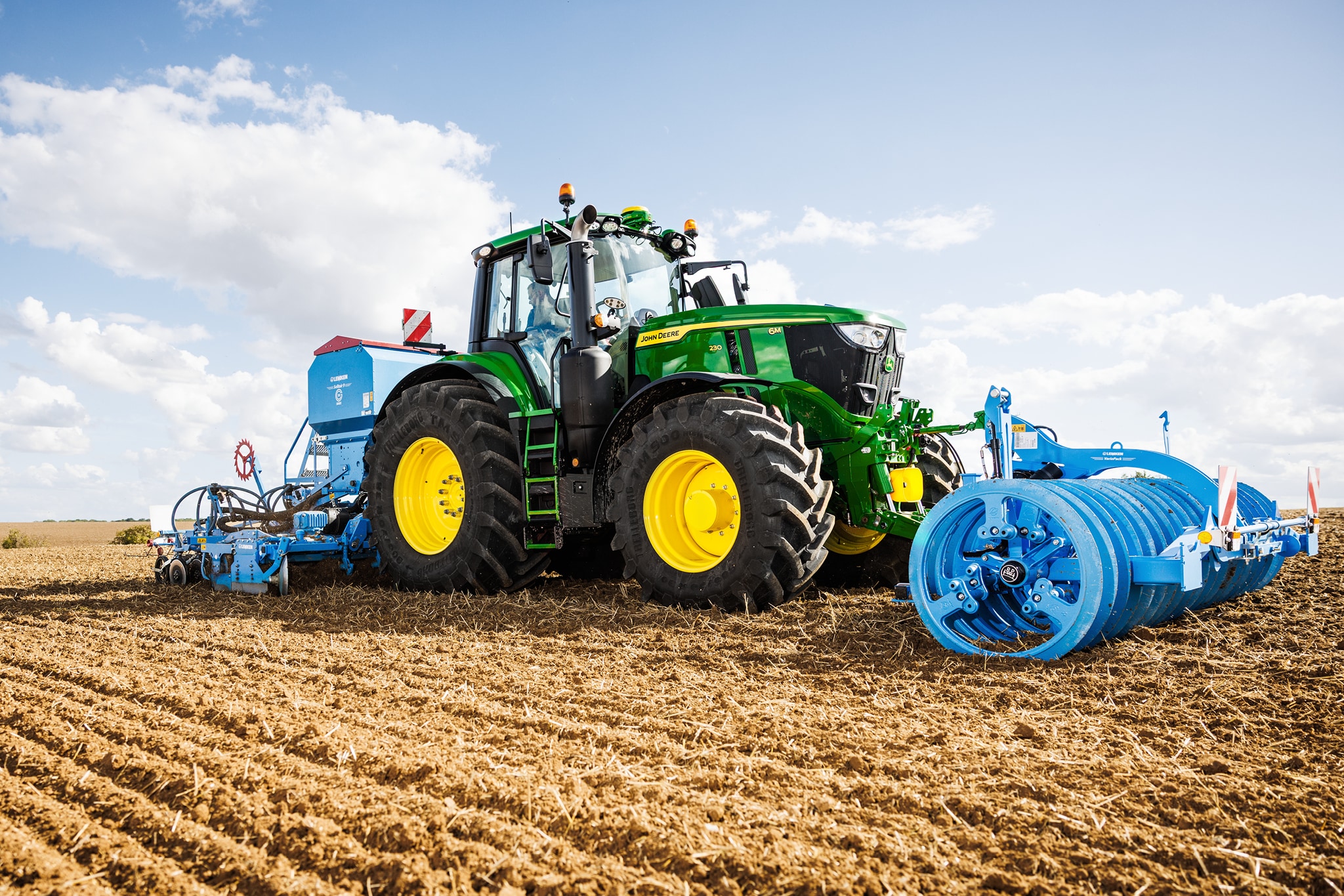 John Deere Traktoren der Serie 6M, die ein Feld mit einem großen Anbaugerät mit blauer Rolle bearbeiten.