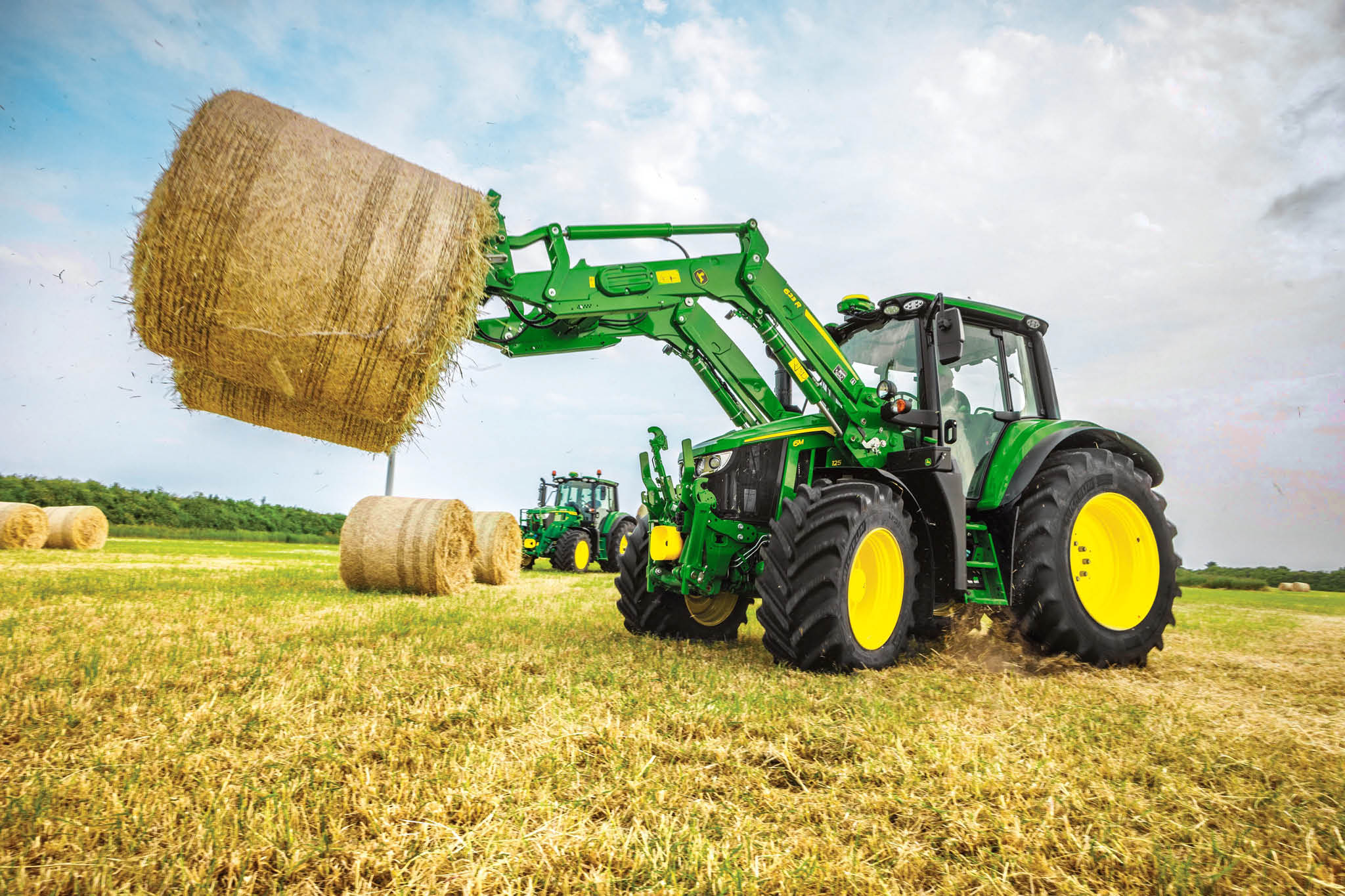 Un tracteur John Deere de la série 6M soulève une grosse balle de foin ronde à l’aide d’un chargeur frontal dans un champ ouvert.
