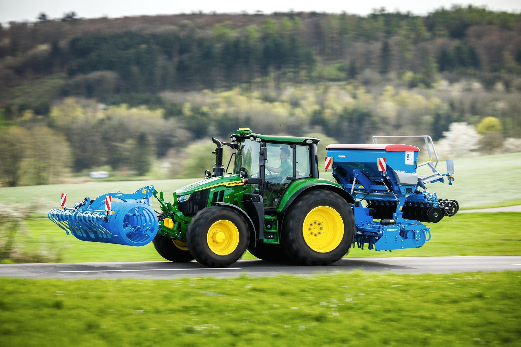 Un tracteur John Deere de la série 6M transportant un équipement monté à l’avant et un semoir à l’arrière sur une route de campagne.