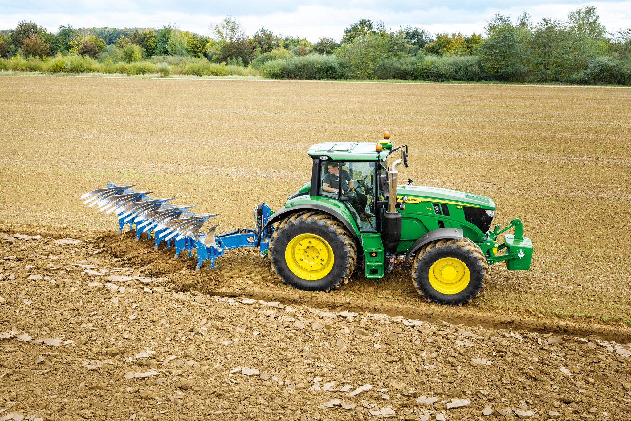 John Deere Traktoren der Serie 6M, die auf einem gepflügten Feld mit einem heckmontierten Bodenbearbeitungsgerät unter bewölktem Himmel arbeiten.