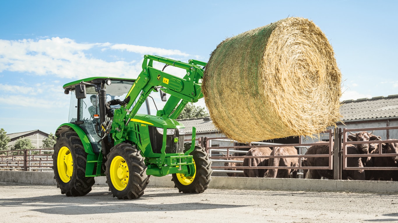 John Deere 5M Traktor auf einem Futterbaubetrieb beim transportieren von Rundballen mit Hilfe eines John Deere Frontladers.