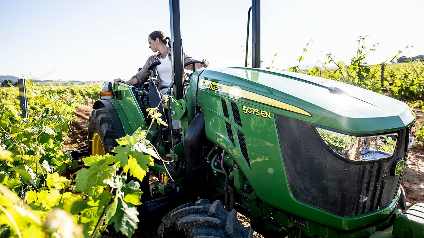 Tracteur John Deere 5075EN dans un vignoble entouré de vignes vertes sous un soleil éclatant.