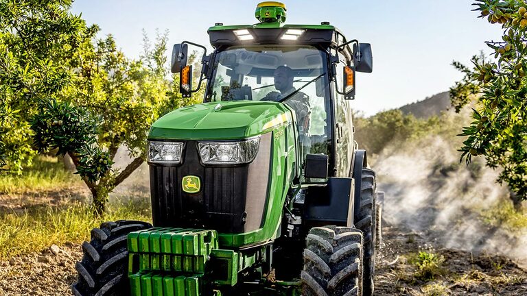 Tracteur John Deere 5075EN vert et jaune traversant un verger baigné de soleil, sur un chemin de terre entouré d’arbres fruitiers.