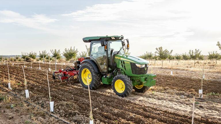 Tracteur John Deere 5075EN cultivant le sol dans un champ ouvert avec ciel dégagé et rangées plantées.