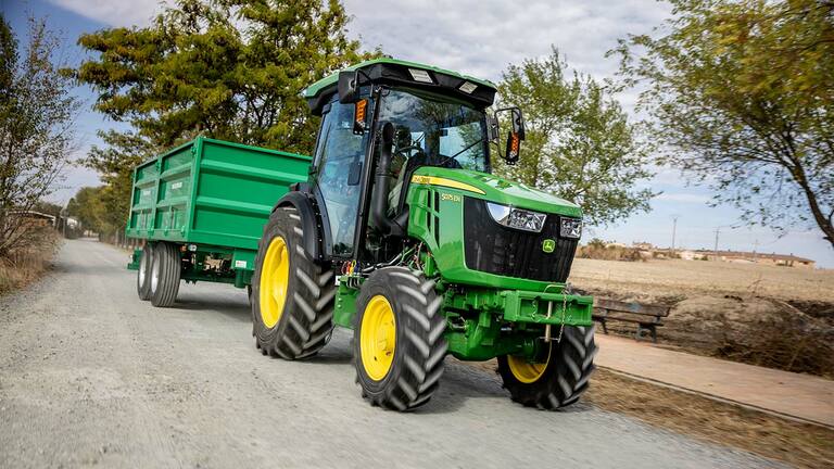 Tracteur John Deere 5075EN tirant une remorque verte sur un chemin rural avec des arbres en arrière-plan.