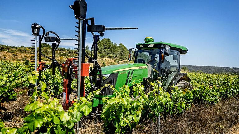Tracteur John Deere 5090EN vert et jaune équipé d’outils de taille dans un vignoble, sous un ciel bleu clair.