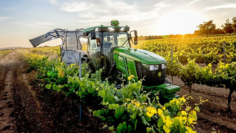 Tracteur John Deere 5105EN vert et jaune pulvérisant les cultures entre les rangs de vigne au coucher du soleil.