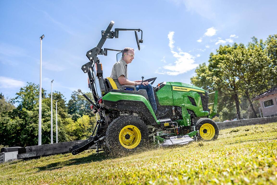 Fahrer, der mit einem John&nbsp;Deere Kompakttraktor und einem Zwischenachsmähwerk einen Grashang mäht
