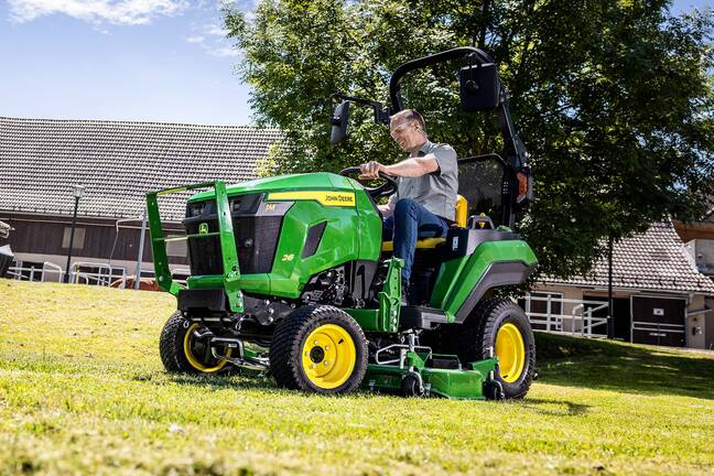 Fahrer, der mit einem John&nbsp;Deere Kompakttraktor und einem Zwischenachsmähwerk einen Grashang mäht