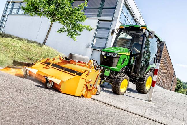 John Deere Kompakttraktor mit geschlossener Kabine und Frontkehrmaschine, der neben einem modernen Gebäude Laub unter Bäumen kehrt